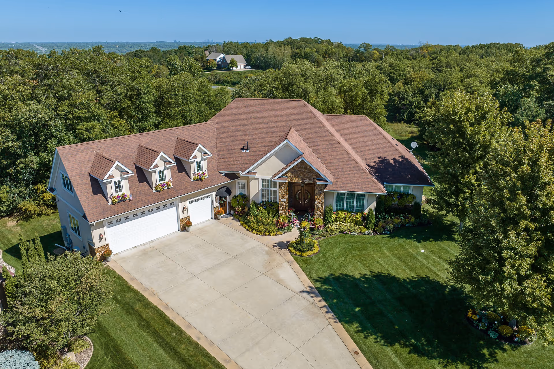 Aerial view of a large single-family house with a brown roof, three dormer windows with flower boxes, a three-car garage, and a well-maintained front yard with green grass, trees, and landscaped flower beds under a clear blue sky.