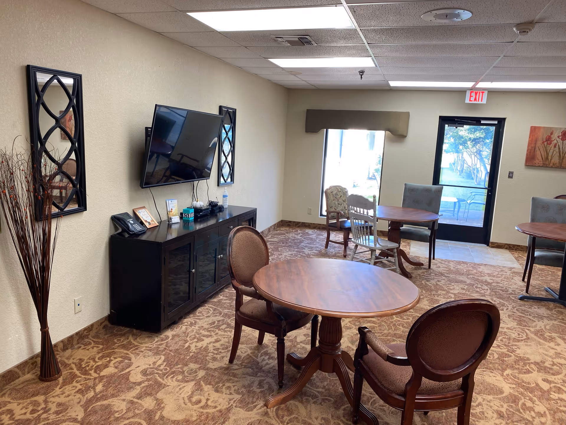 A senior living common room with round wooden tables and chairs, a wall-mounted TV above a cabinet, and an exit door with windows to the outside.