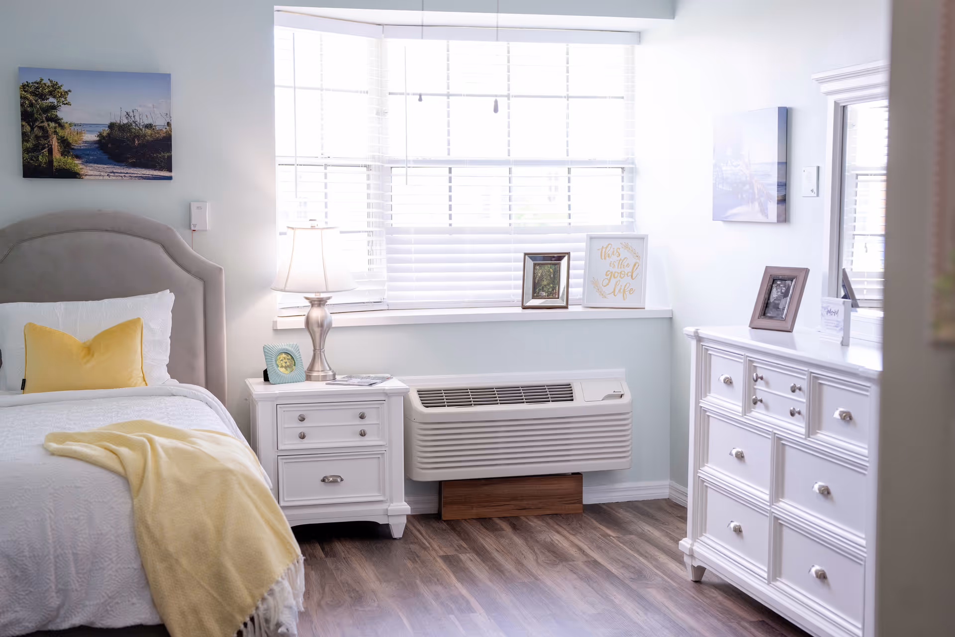 A bright and cozy bedroom with a bed featuring a gray upholstered headboard, white bedding, and a yellow throw blanket and pillow. Next to the bed is a white nightstand with a silver lamp and a small framed photo. A large window with white blinds lets in natural light. Below the window is a wall-mounted air conditioning unit. On the right side of the room is a white dresser with multiple drawers and a mirror above it, decorated with framed photos and a small sign that reads 'this is the good life'. The floor is wood with a natural finish.