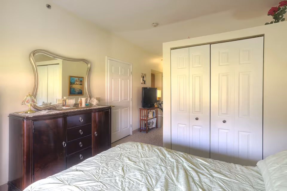 Bright bedroom with a bed in the foreground, a dark wooden dresser topped with a decorative mirror and accessories, and white closet doors and a TV in the background.