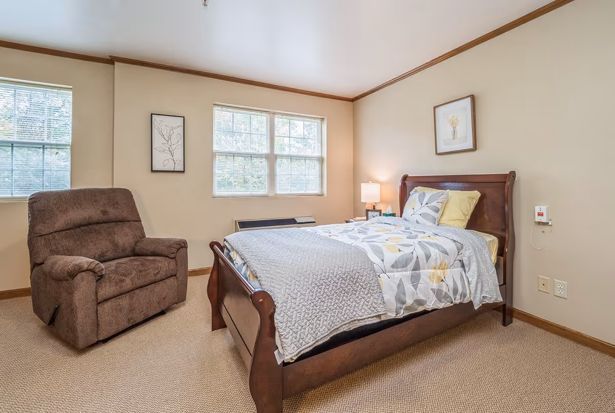 A cozy bedroom with a wooden bed frame, a bed made with floral-patterned bedding and pillows, a brown recliner chair, two windows with blinds, a nightstand with a lamp, and framed artwork on the walls.