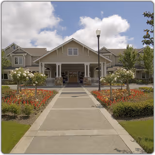 Front exterior view of Aegis Living Corte Madera facility with a paved walkway leading to the entrance, surrounded by landscaped flower beds and greenery under a partly cloudy sky.