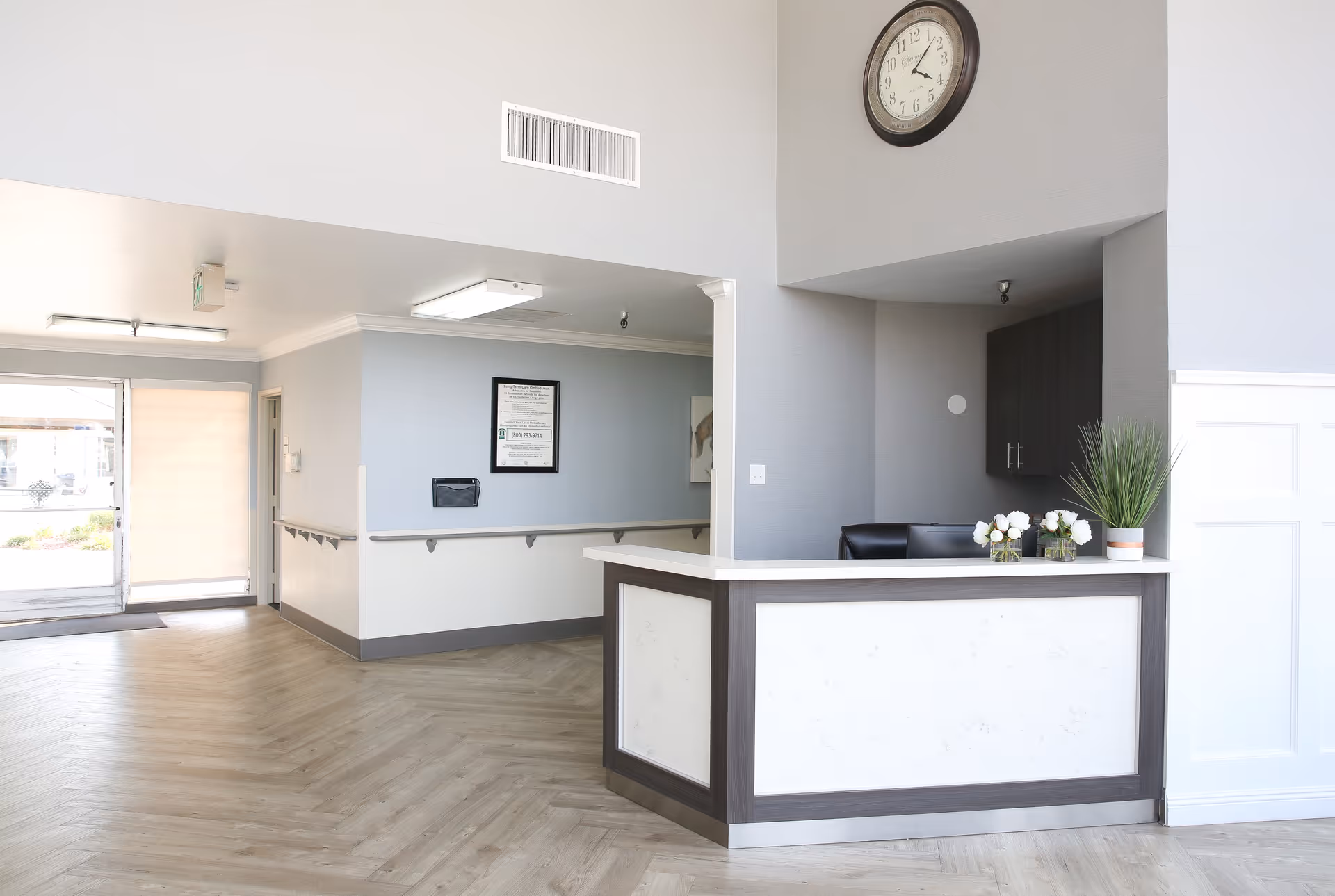 Reception desk and lobby area with a wall clock, potted plants, and an entrance door.
