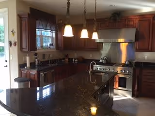 A modern kitchen with dark granite countertops, wooden cabinets, a large stainless steel stove with an overhead vent, a sink on the island countertop, and three pendant lights hanging above the island. There is a window with a valance above the sink and a door to the left side.