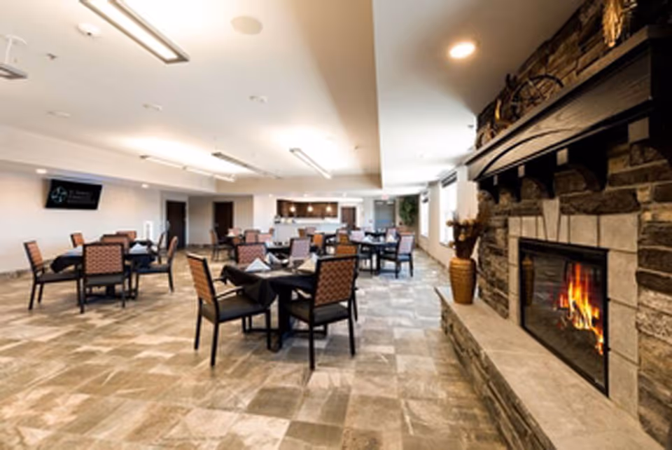 A spacious dining room with several tables and chairs arranged neatly. Each table is set with black tablecloths and folded napkins. On the right side, there is a stone fireplace with a fire burning, and decorative items on the mantel. The room has tiled flooring, bright overhead lighting, and windows along the right wall letting in natural light.