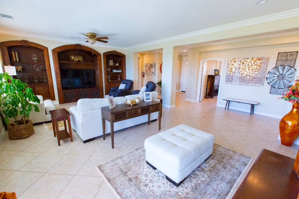 A spacious and well-lit living room in a senior living facility featuring white sofas, a large ottoman on a patterned rug, two dark armchairs, a wooden console table with decorative items, built-in wooden shelves with ornaments, a flat-screen TV, a large potted plant, and wall art including a metallic windmill and a tree painting.