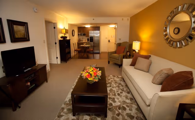 A cozy living room with a beige sofa adorned with multiple cushions, a patterned rug, and a dark wooden coffee table with a colorful flower arrangement. To the left is a TV on a wooden stand, and in the background, there is a small dining area with a round table and two chairs, as well as a kitchen with stainless steel appliances. The walls are painted in warm tones, and there is a decorative round mirror above the sofa.
