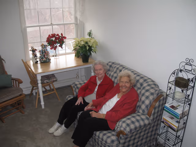 Two elderly women sitting on a blue and white checkered couch in a cozy room with a window behind them. A wooden table with poinsettia plants and holiday decorations is near the window. There is a wooden chair and a metal shelf with books and decorative items on the right side.