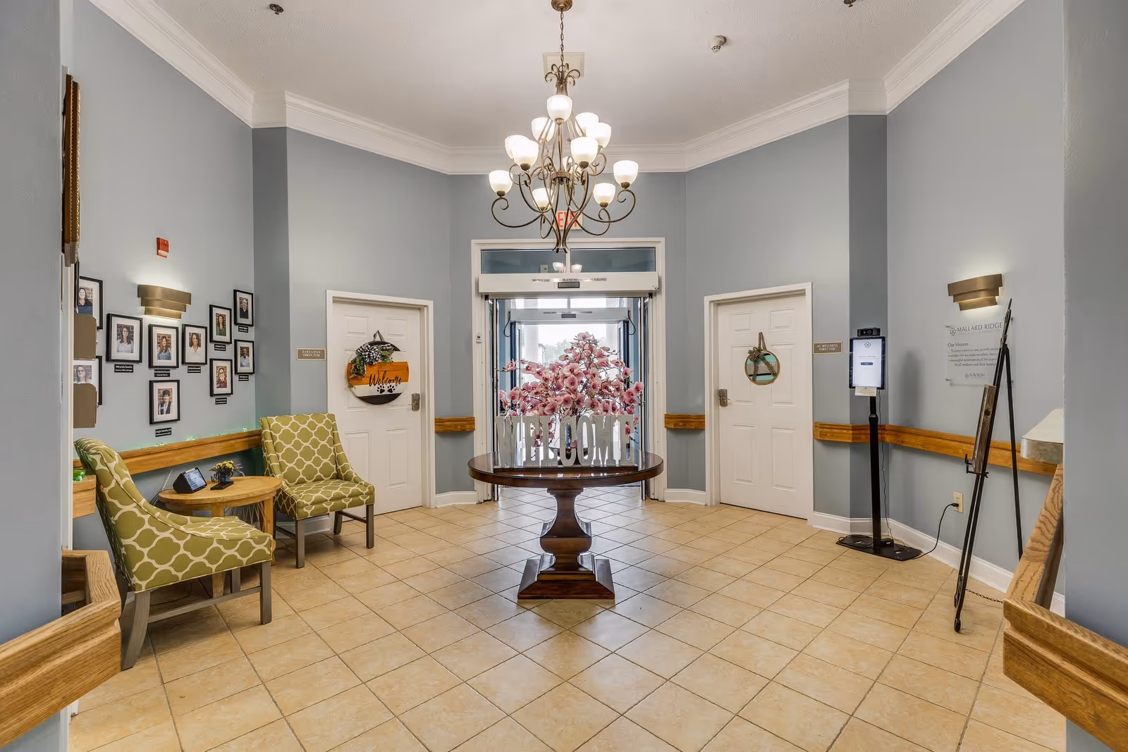 A welcoming lobby area with light blue walls, beige tiled floor, and a chandelier hanging from the ceiling. In the center, there is a round wooden table with a floral arrangement and a decorative 'WELCOME' sign. Two green patterned chairs and a small round wooden table are placed to the left. There are framed photos on the left wall and two white doors on either side of the entrance door. A hand sanitizer station and a sign with the facility's mission are on the right wall.