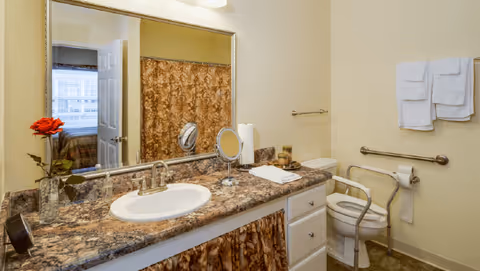 A bathroom with a granite countertop sink, a large mirror above the sink, a toilet with safety rails, a towel rack with white towels, and a shower with a patterned curtain. A red flower in a vase is placed on the countertop, and a bedroom is partially visible through an open door.