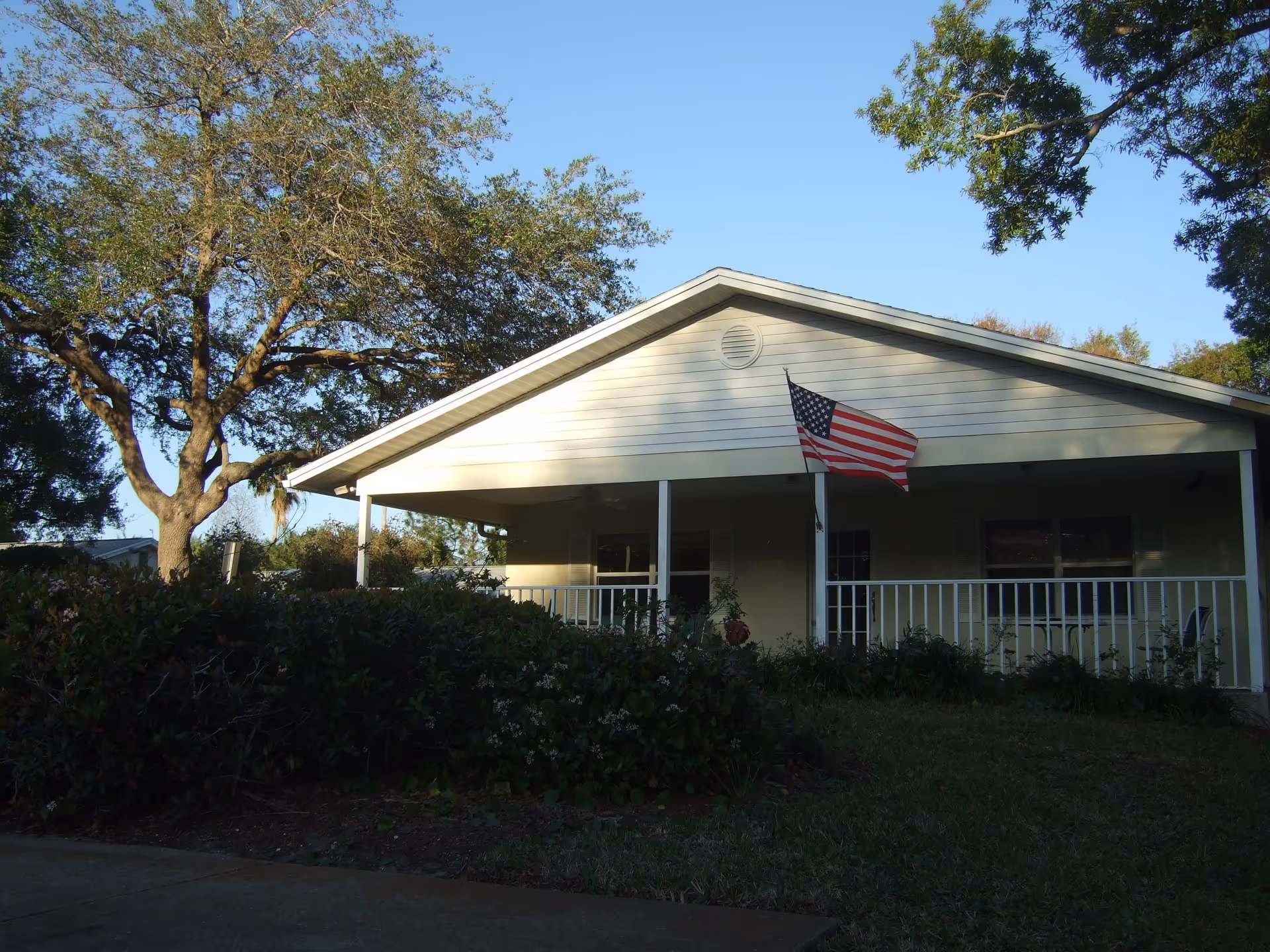 Single-story building with a covered porch, white railing, and an American flag mounted near the entrance. The building is surrounded by trees and bushes under a clear blue sky.