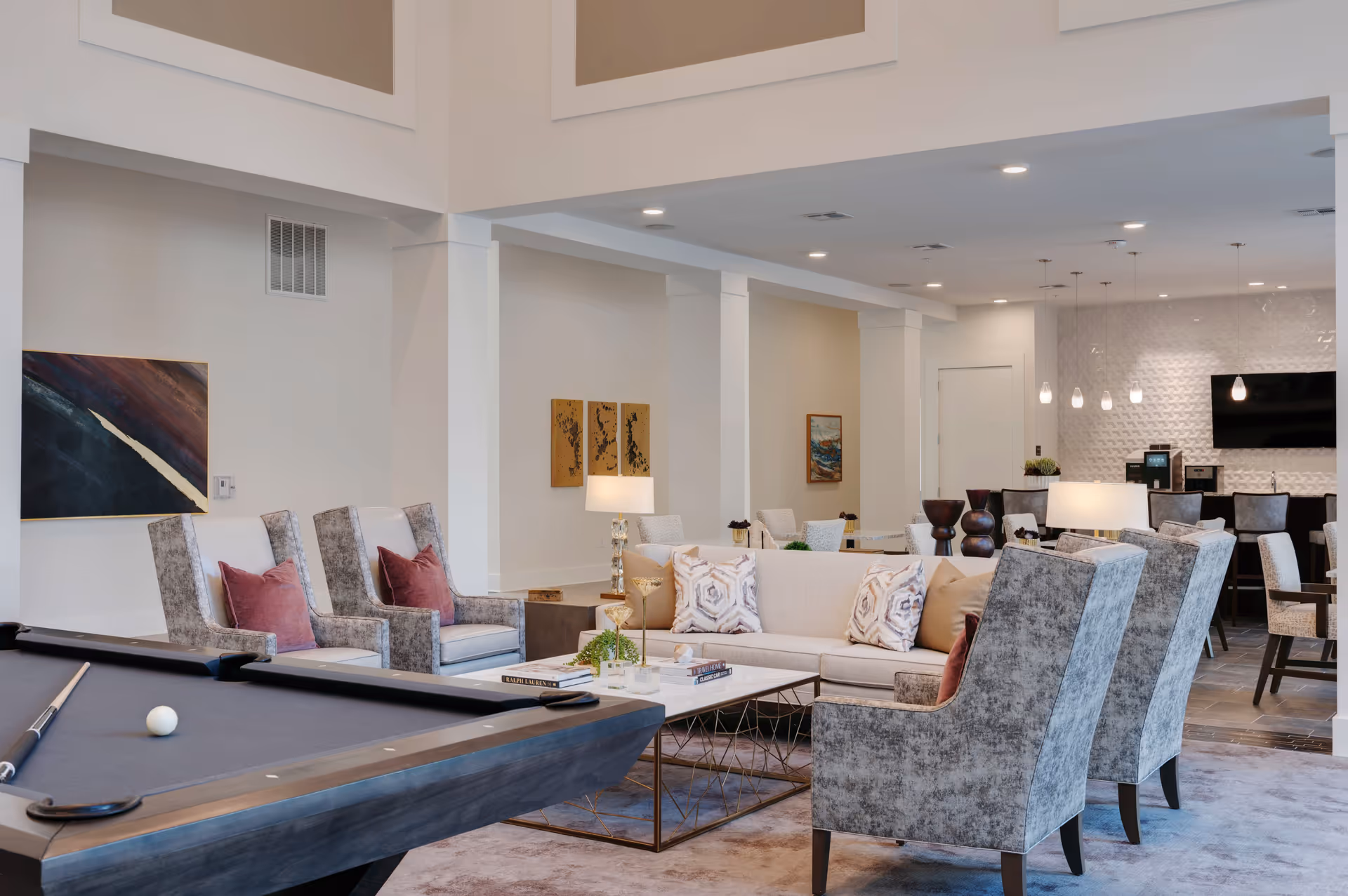 A spacious and modern common area featuring a pool table in the foreground, a beige sofa with patterned pillows, several upholstered armchairs with red cushions, a coffee table with decorative items, and a dining area with tables and chairs in the background. The room has high ceilings, recessed lighting, and contemporary artwork on the walls.