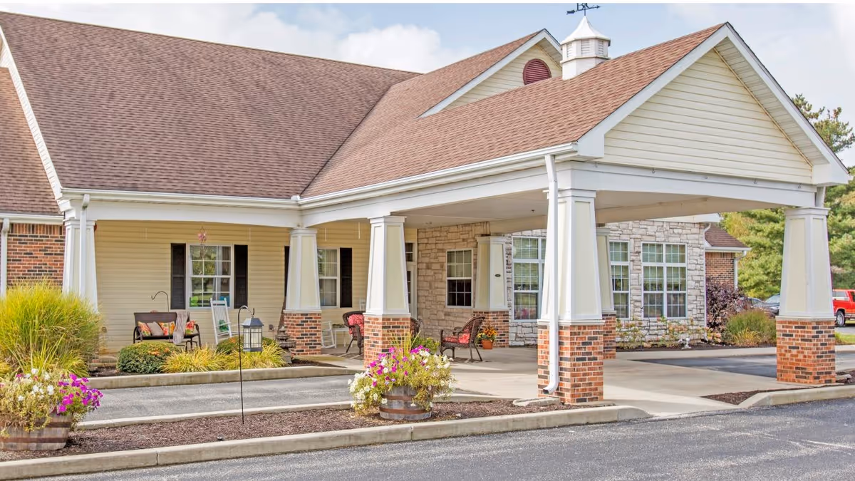 Covered front entrance of a senior living facility with brick columns, seating, and flower planters.