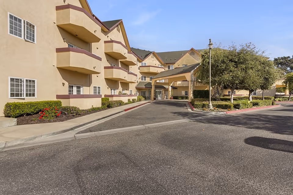 Exterior view of a multi-story senior living facility with beige walls and maroon trim. The building features balconies, windows, and a covered entrance driveway. There are landscaped bushes and trees along the sidewalk and driveway under a clear blue sky.