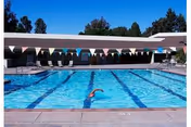 Outdoor rectangular swimming pool with lane markers and colorful pennant flags overhead, a swimmer in a lane, and a low building with lounge chairs in the background.