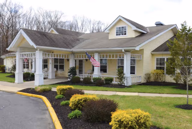 Exterior view of a single-story yellow building with white trim, a covered entrance supported by white columns, an American flag hanging near the entrance, and landscaped bushes and grass surrounding the walkway.