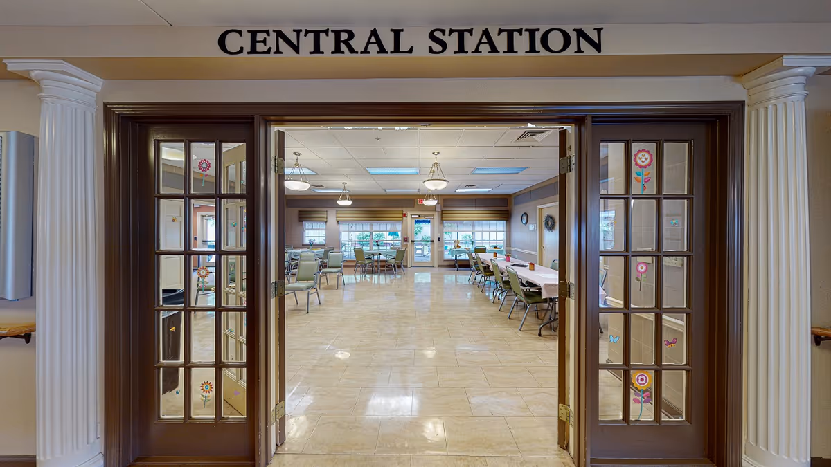 View through double glass doors with flower and butterfly decals into a large, well-lit room with multiple tables and chairs arranged for dining or activities. The room has beige tiled floors, several windows with blinds, and ceiling lights. Above the doors is a sign that reads 'CENTRAL STATION'.