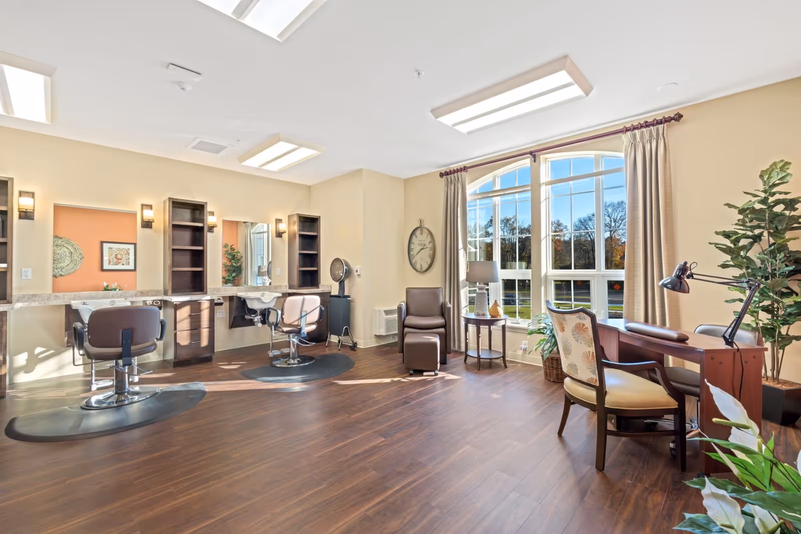 A bright and spacious salon area in a senior living facility with two salon chairs in front of mirrors and shelves, a comfortable armchair with an ottoman, a side table with a lamp, and a desk with a chair near large arched windows letting in natural light. The room has wooden flooring and beige walls with plants adding greenery.
