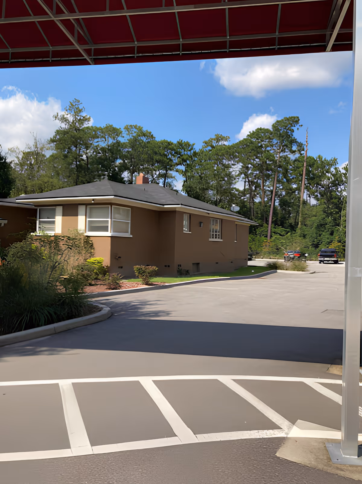 View of a single-story brown building with white-framed windows surrounded by greenery and trees under a blue sky with some clouds. The image is taken from a paved area with white painted lines, possibly a parking or driveway area, with a red awning partially visible at the top.