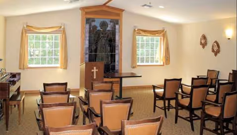 Chapel-style room in a senior facility with rows of chairs, lectern, cross, and windows.