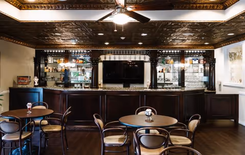 Interior view of a senior living facility lounge area with a dark wood bar counter, glass shelves displaying various bottles and glasses, a large flat-screen TV mounted on the wall behind the bar, round tables with chairs arranged in front, and an ornate decorative ceiling with a ceiling fan and light fixture.