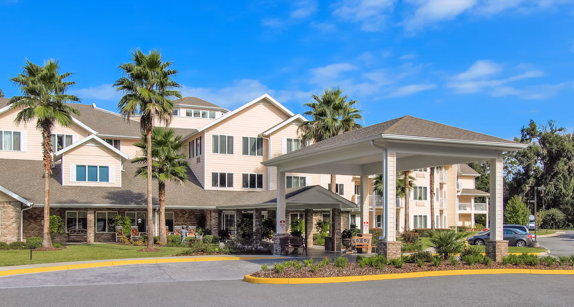 Exterior view of a multi-story senior living facility with beige siding and stone accents, surrounded by palm trees and landscaping. There is a covered entrance driveway with seating areas and a clear blue sky in the background.