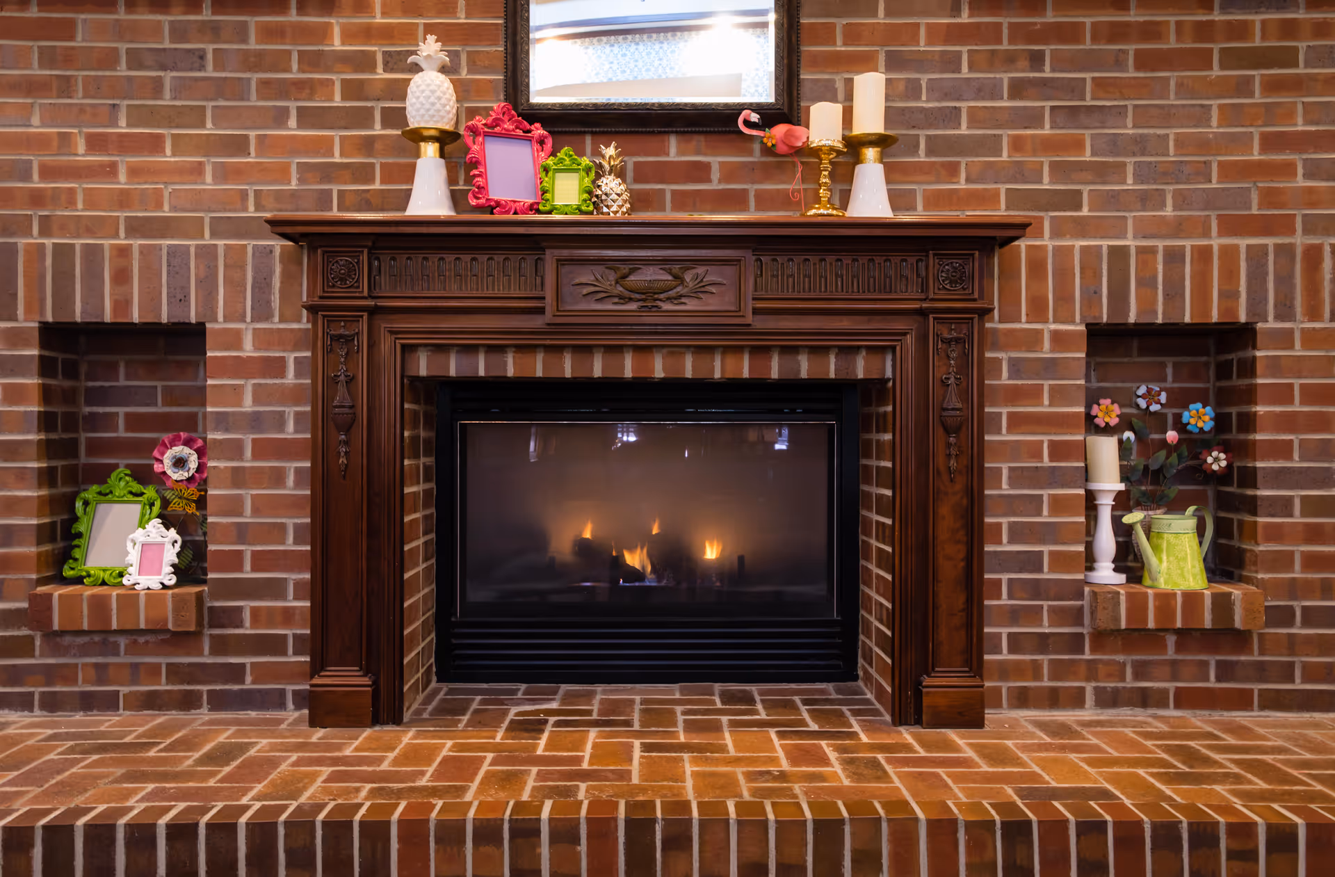 A cozy brick fireplace with a dark wooden mantel. On the mantel are decorative items including two white candle holders with candles, a small golden pineapple, and colorful picture frames. The fireplace is lit with a warm flame. On either side of the fireplace are built-in brick shelves holding additional decorative items such as picture frames, a watering can, and flower decorations.