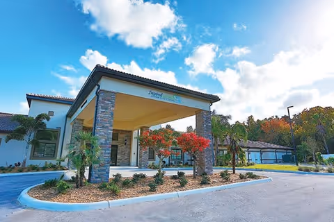 Front exterior view of Inspired Living Sun City Center facility with a covered entrance supported by stone pillars, landscaped plants and trees, and a clear blue sky with some clouds.