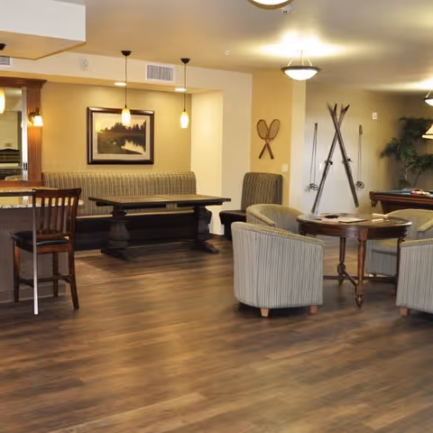 A cozy common area with wooden flooring featuring a round table surrounded by four cushioned armchairs. On the wall behind, there are decorative vintage wooden skis and tennis rackets. To the left, there is a built-in bench with a dark wooden table and two pendant lights hanging above. A framed landscape painting is mounted on the wall above the bench. The room has warm lighting and a comfortable, inviting atmosphere.