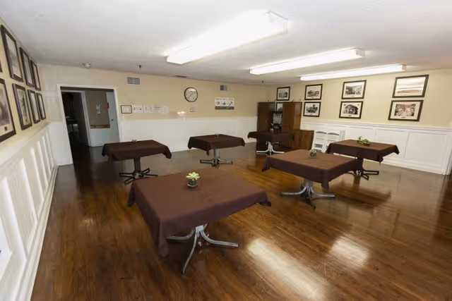 A dining room with six square tables covered with brown tablecloths, each table has a small plant centerpiece. The room has wooden floors, cream-colored walls with white wainscoting, and framed pictures hanging on the walls. There is a clock and some notices on one wall, and a wooden cabinet in the corner.