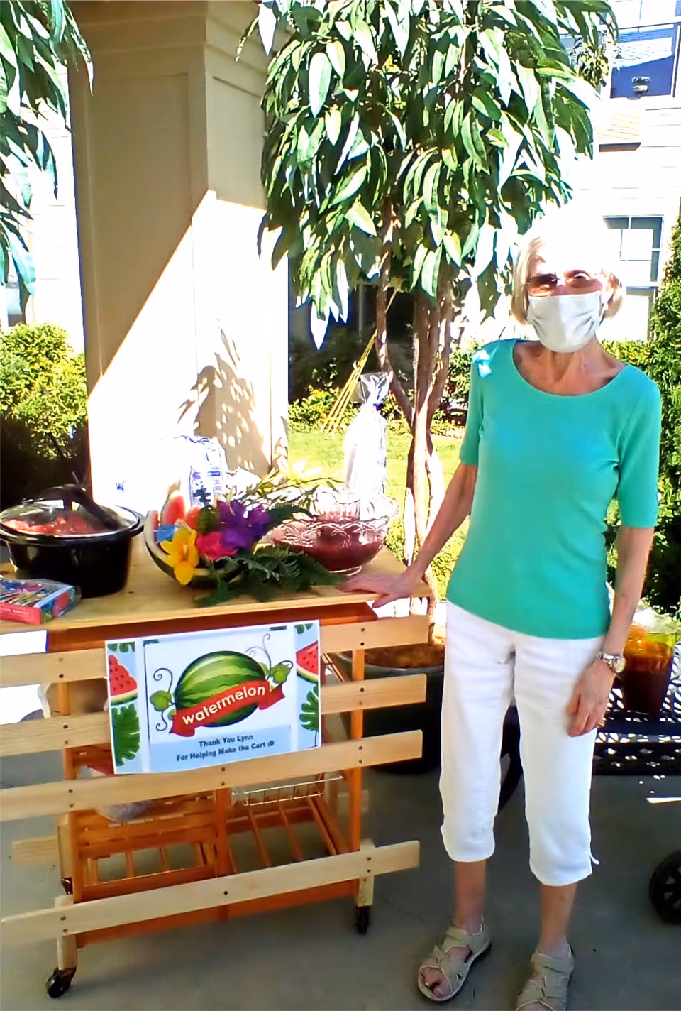 An elderly woman wearing a green top, white pants, sandals, sunglasses, and a face mask stands next to a wooden cart with a watermelon-themed sign. The cart holds a bowl of watermelon slices, a crockpot, a pitcher of red drink, and a floral arrangement. The scene is outdoors with greenery and a building in the background.