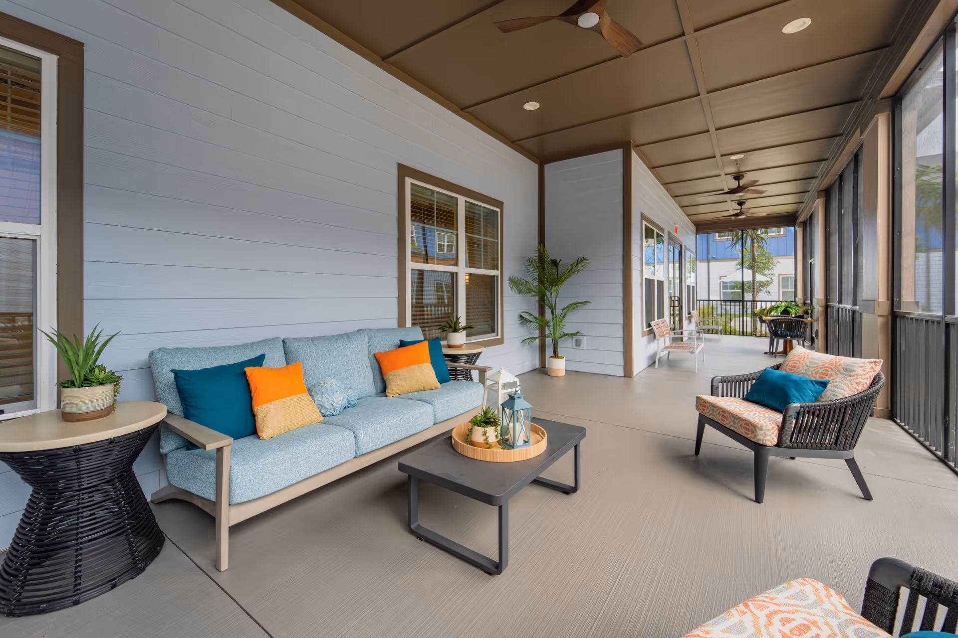 A spacious screened-in porch area with comfortable seating including a light blue sofa with colorful cushions, a black wicker chair with patterned cushions, and a black coffee table with decorative items. The porch has ceiling fans, potted plants, and large windows looking out to an outdoor area.