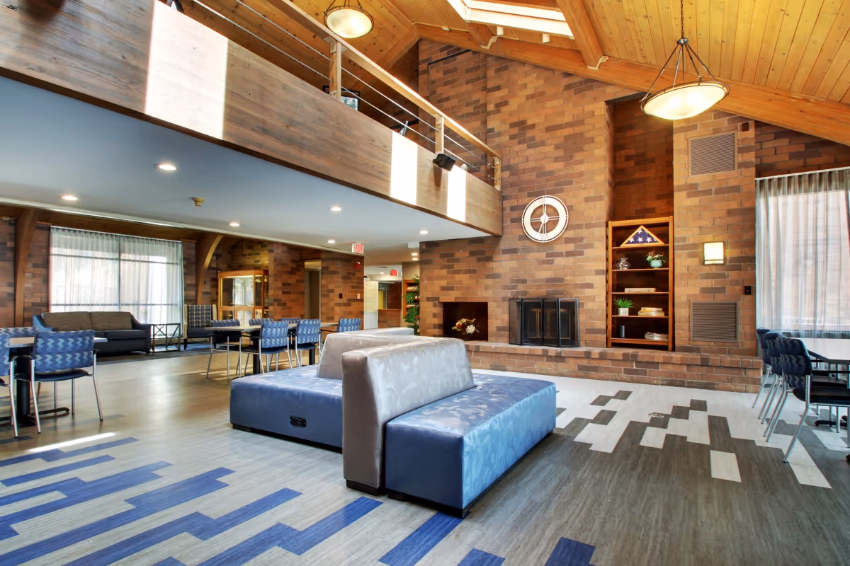 A spacious senior living facility common area with a high wooden ceiling and brick walls. The room features a large fireplace with a clock above it, built-in shelves with decorative items, and modern seating including blue and gray cushioned benches and chairs around tables. Large windows with sheer curtains allow natural light to fill the space.