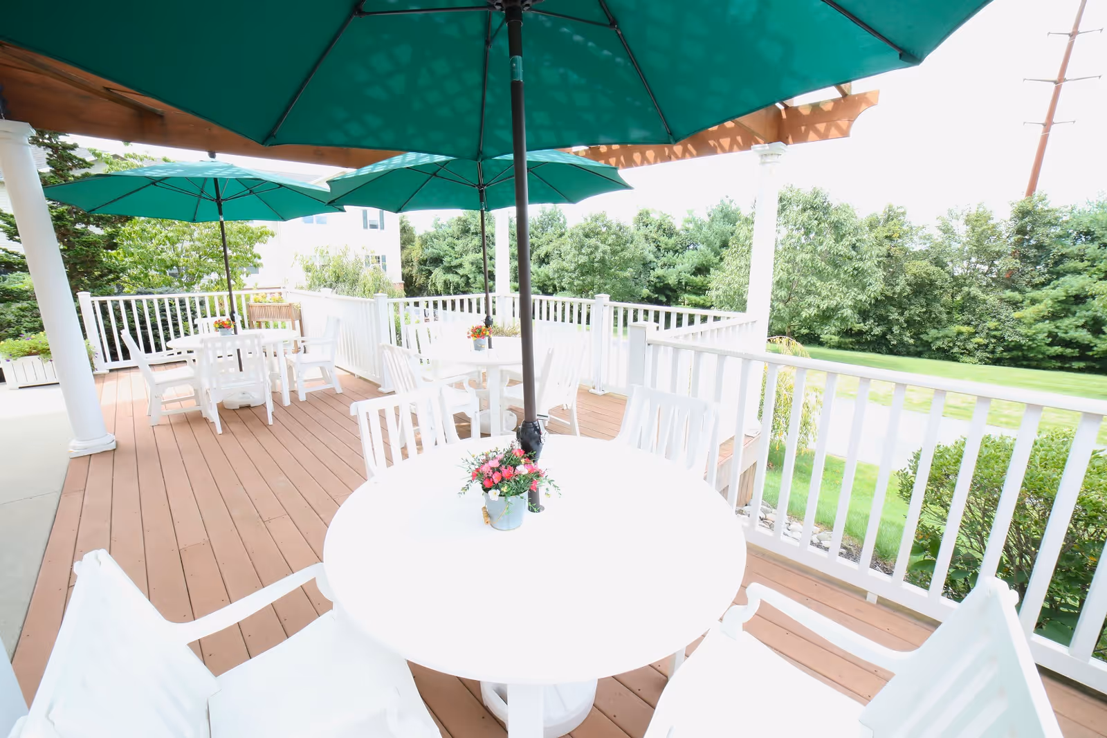Outdoor patio area with white round tables and white chairs under large green umbrellas. The patio has a wooden floor and white railing, surrounded by greenery and trees in the background.