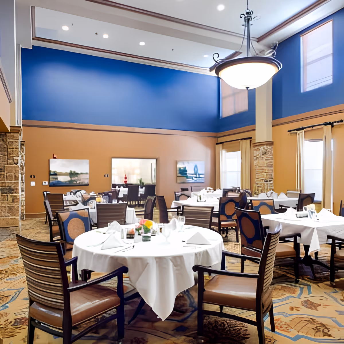 A spacious dining room with several round and rectangular tables covered with white tablecloths, set with glassware and napkins. The room features high ceilings, large windows with beige curtains, blue and tan walls, and a large hanging light fixture. There are multiple chairs around each table and decorative paintings on the walls.