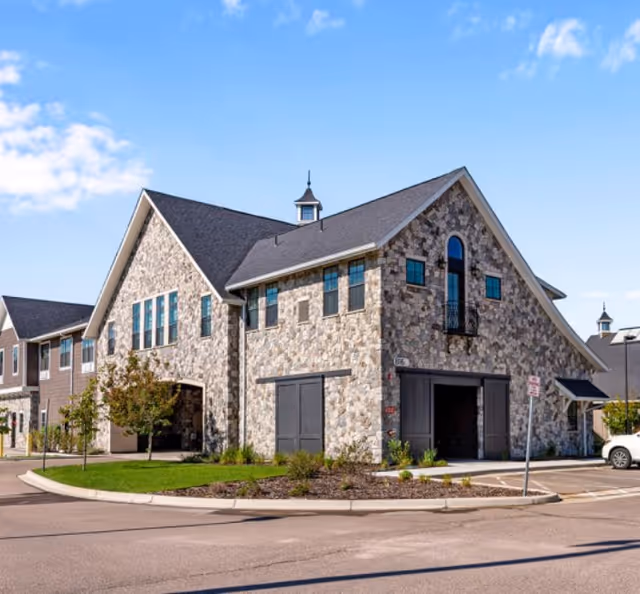 Stone-faced, two-story building with peaked roofs, garage bays and landscaped curbside under a blue sky.