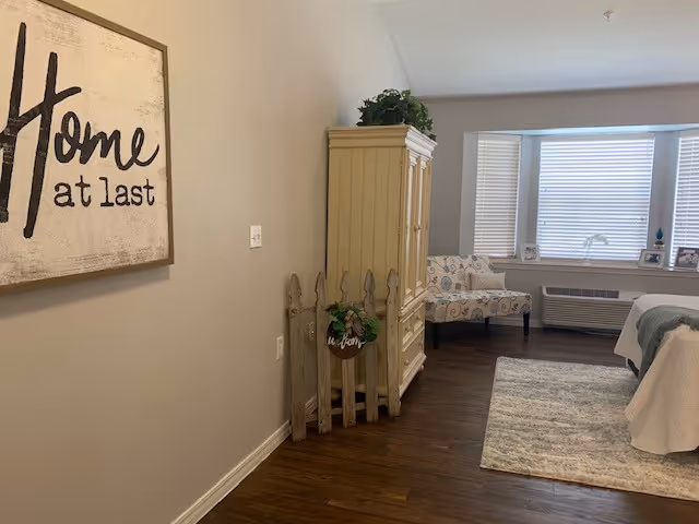 A cozy interior room with wooden flooring, a beige wall with a framed sign that says 'Home at last', a cream-colored cabinet with a green plant on top, a small decorative wooden fence with a plant, a patterned armchair near a window with blinds, a heating/cooling unit beneath the window, and a bed partially visible on the right with a white and gray blanket on a light-colored rug.