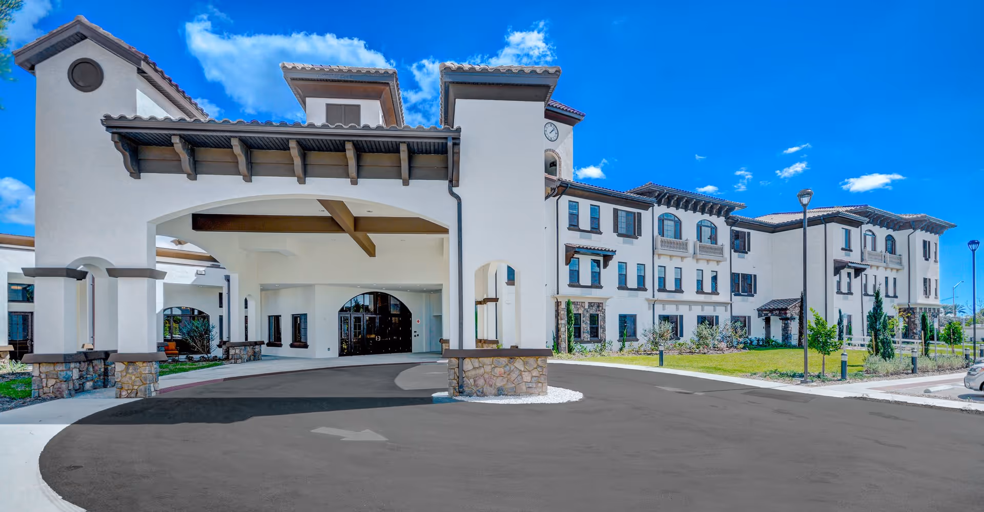 Front exterior view of Tuscan Gardens Of Venetia Bay, a large multi-story building with a covered entrance, stone accents, and a clock tower under a bright blue sky with some clouds.