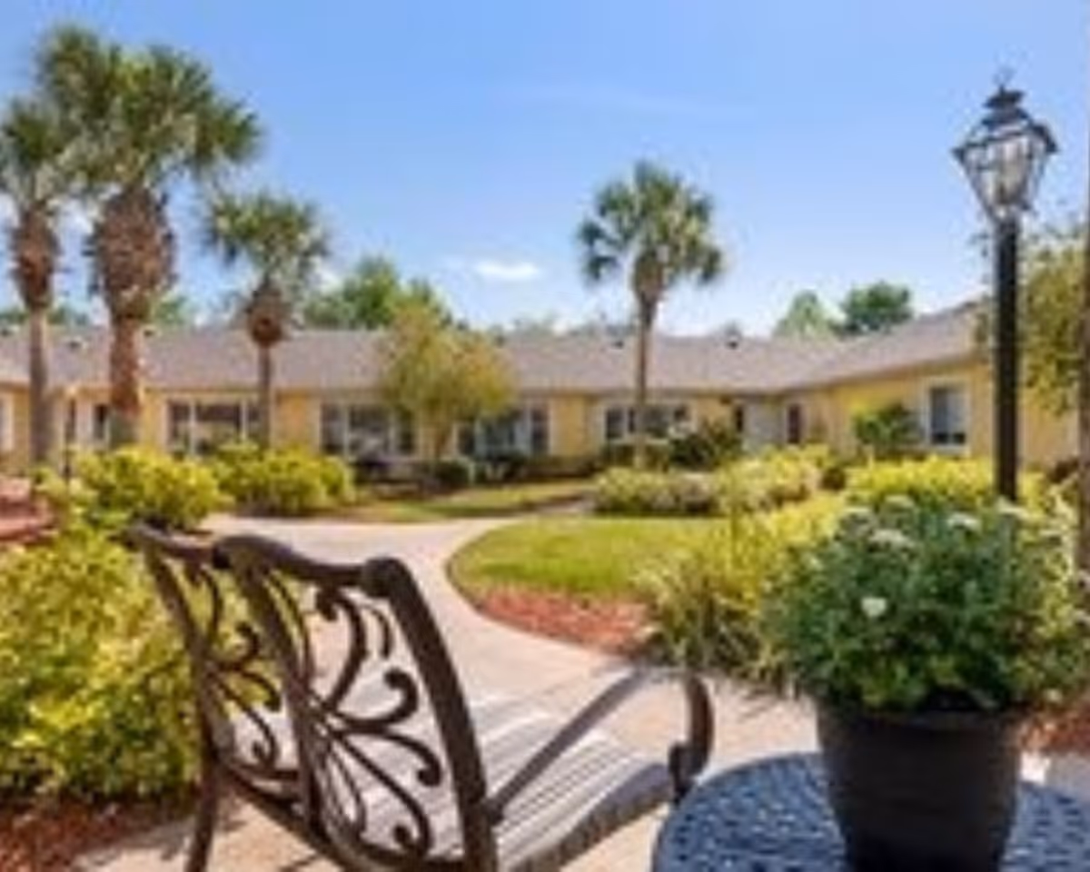 Outdoor courtyard area with a decorative metal bench and a round table holding a potted plant. The courtyard is surrounded by well-maintained greenery, palm trees, and a single-story building under a clear blue sky.
