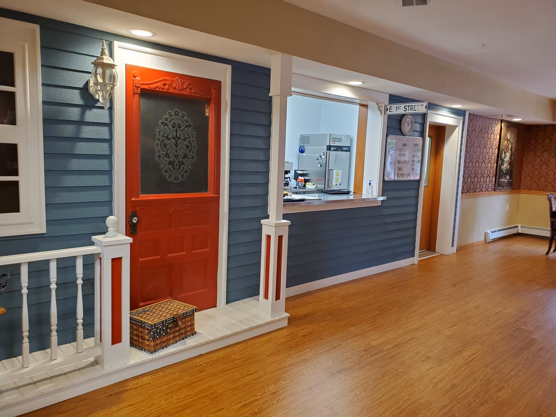 Indoor hallway area designed to look like an outdoor porch with a blue siding wall, a red door with decorative glass, white railing, and a small basket on the porch floor. To the right, there is a serving window with a menu board above it and a clock, next to an elevator door. The floor is wood and the far wall has patterned wallpaper with a framed floral painting.