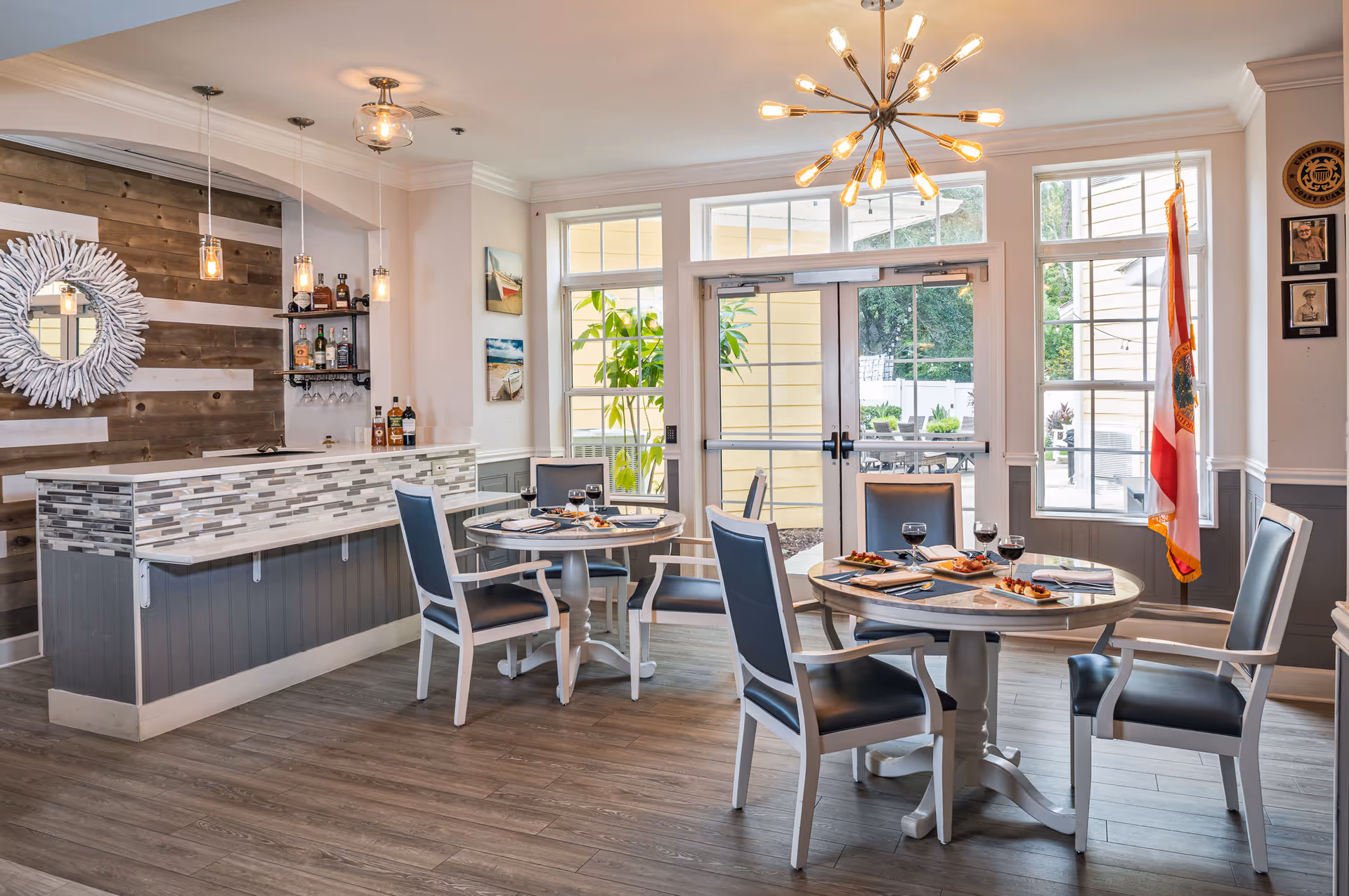 A bright dining area with two round tables set with plates of food and glasses of red wine. The room features modern pendant and chandelier lighting, a small bar with bottles and glasses, large windows and glass doors leading outside, and a Florida state flag in the corner.