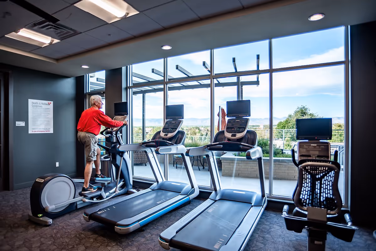 An elderly man in a red shirt and shorts is using an elliptical machine in a fitness room with large windows overlooking an outdoor patio and scenic landscape. The room also contains two treadmills and a stationary bike, each equipped with individual screens.