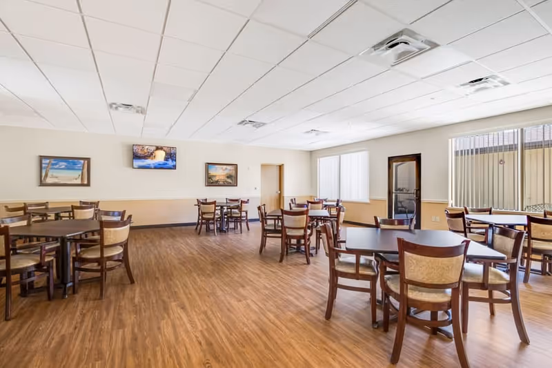 A spacious dining room with multiple wooden tables and chairs arranged neatly on a wood-patterned floor. The room has a white ceiling with recessed lighting and air vents. There are three framed pictures on the far wall and large windows on the right side letting in natural light. A door is visible on the far wall and another door with a window is on the right side.