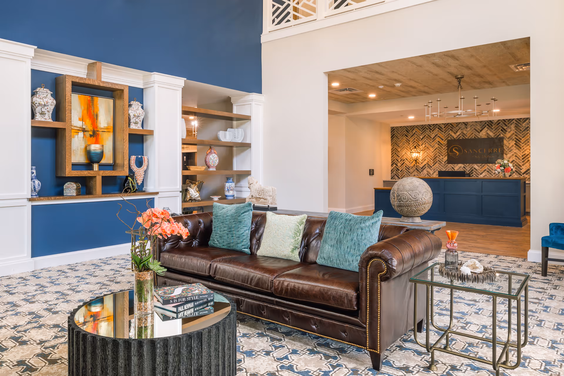 Bright furnished lobby with a brown leather sofa, patterned rug, coffee tables, decorative shelving, and a reception desk in the background.