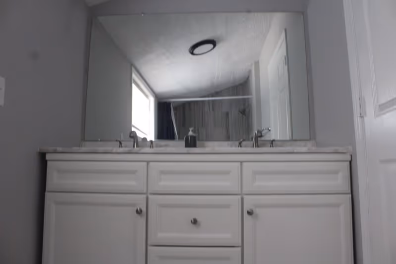 A bathroom vanity with a white countertop and two faucets. Above the vanity is a large mirror reflecting a window and a shower curtain. The walls are painted gray and there is a white door to the right.
