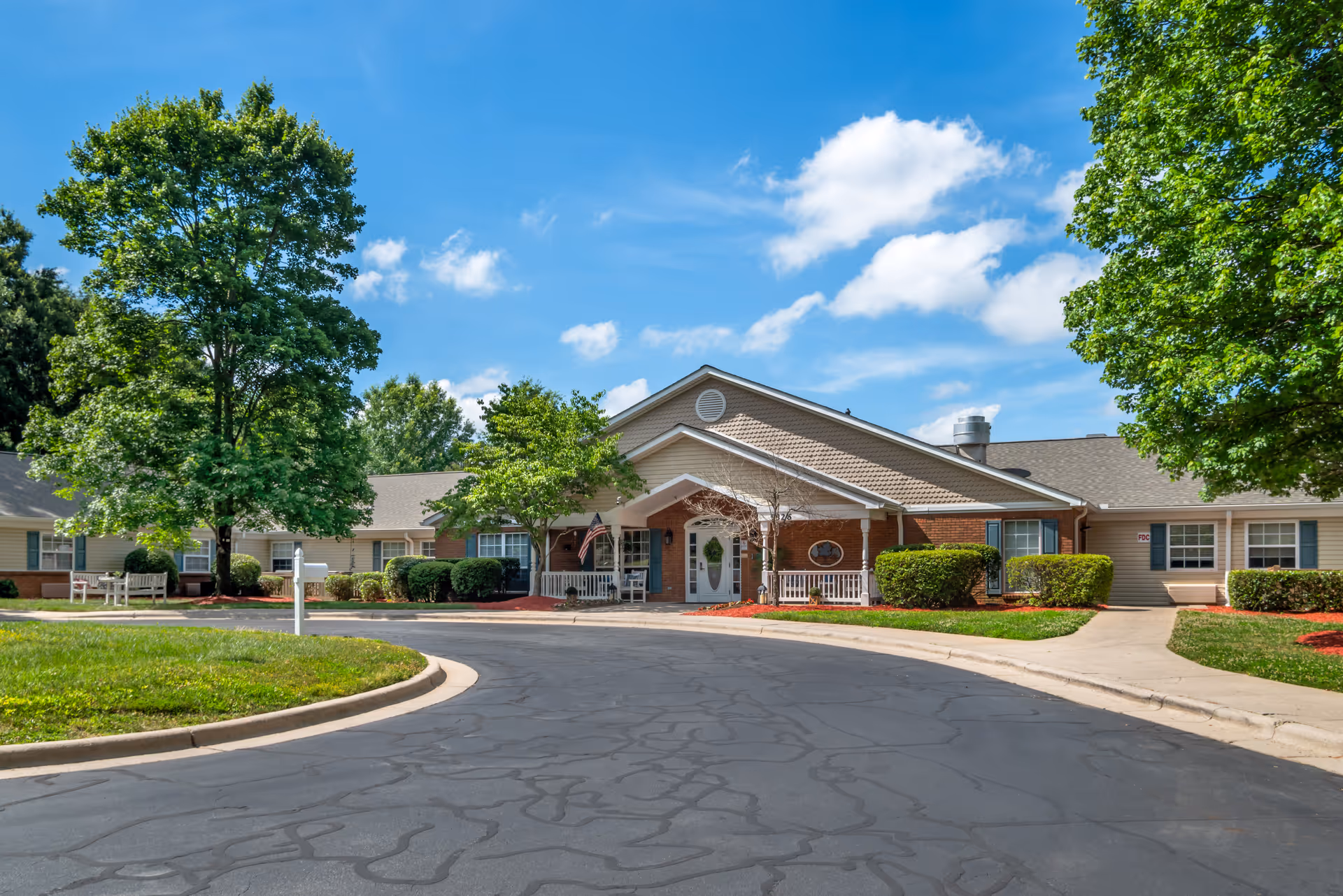 Single-story brick and siding senior living building with a covered entrance, landscaped shrubs and trees, and a circular driveway under a blue sky.