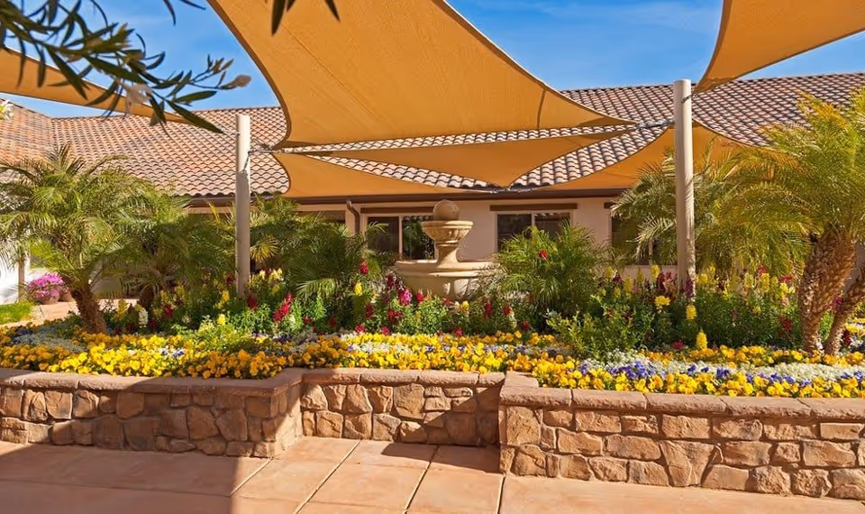 A sunny outdoor garden area with a stone raised flower bed filled with colorful flowers and green plants. There are palm trees and a decorative water fountain in the center. Beige shade sails are stretched overhead, and a building with a tiled roof is visible in the background under a clear blue sky.