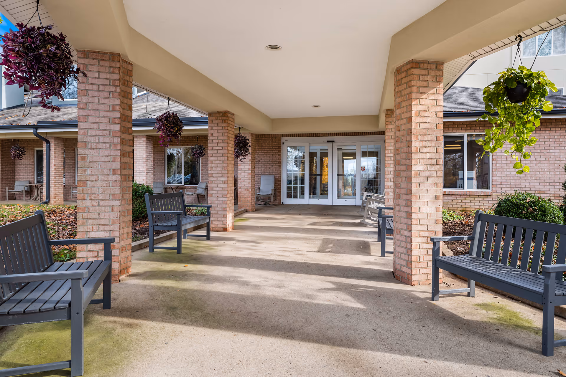 Covered outdoor seating area at the entrance of a senior living facility with brick pillars, hanging plants, and several benches along the walkway leading to glass double doors.