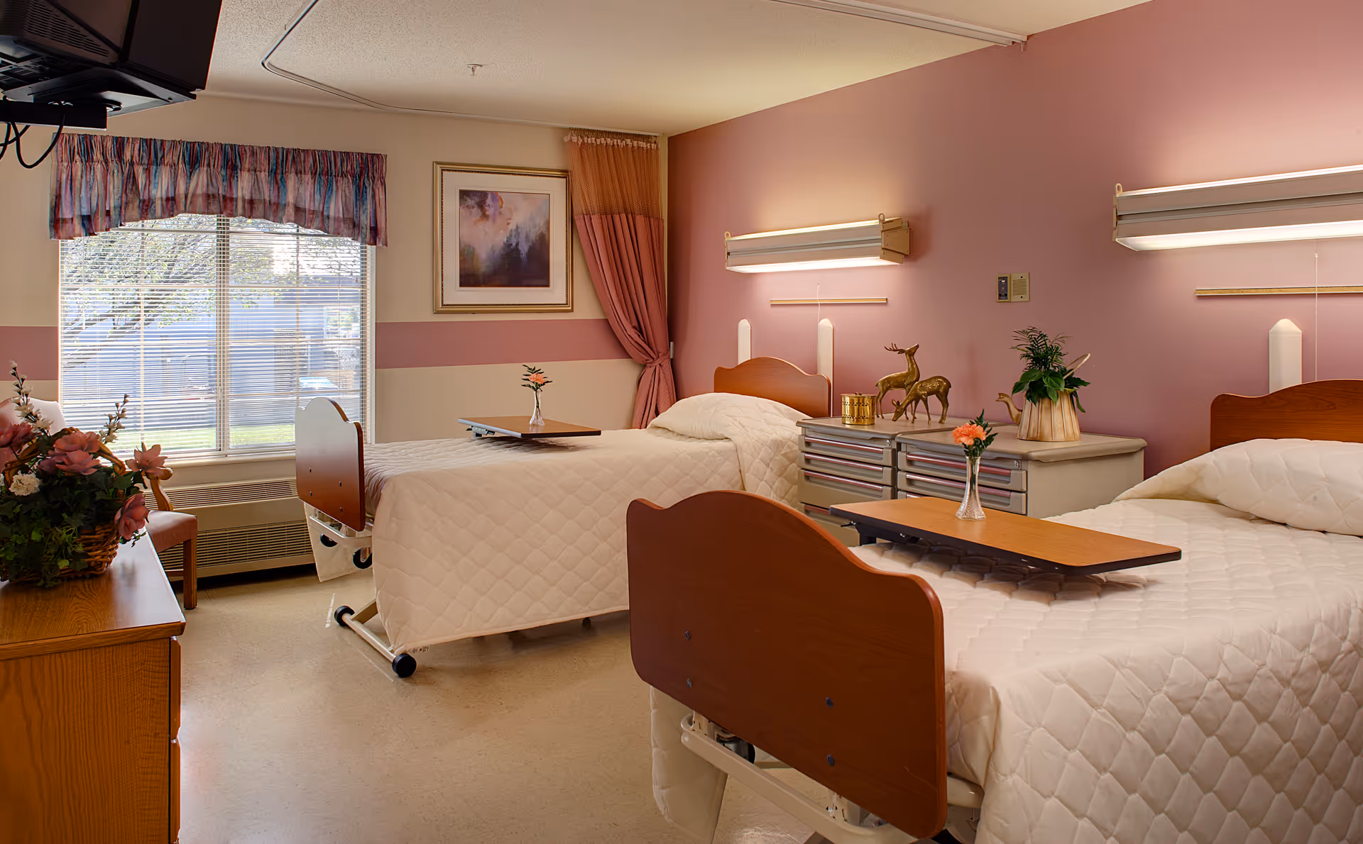 A shared senior living bedroom with two hospital-style beds, bedside tables, and a window with curtains.