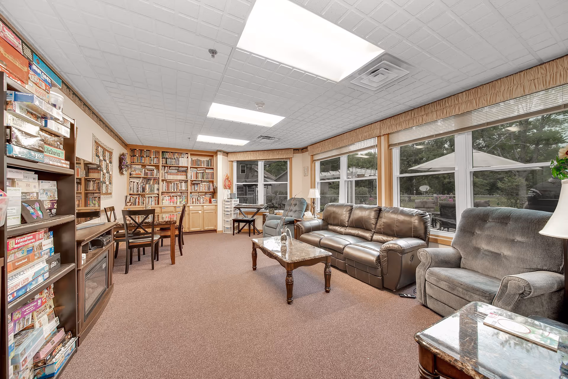 A cozy common room with large windows letting in natural light, featuring a brown leather sofa, a gray armchair, a marble-top coffee table, a bookshelf filled with books, a game shelf with board games, a wooden table with chairs, and a keyboard on a stand near the window.